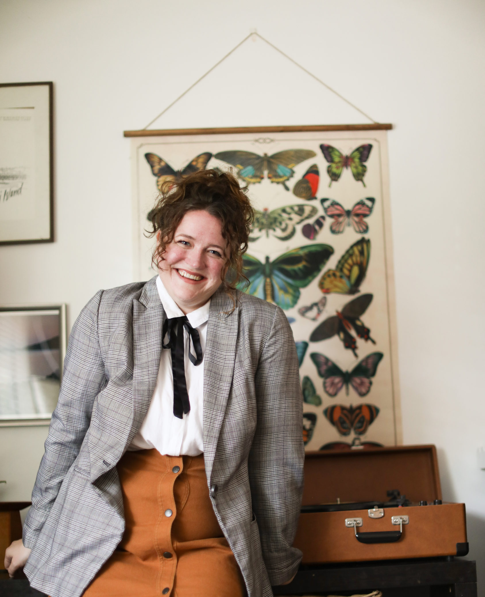 A young woman in a blazer and a white button down with a black tie smiles at the cmaera in front of a record player and a poster with butterflies